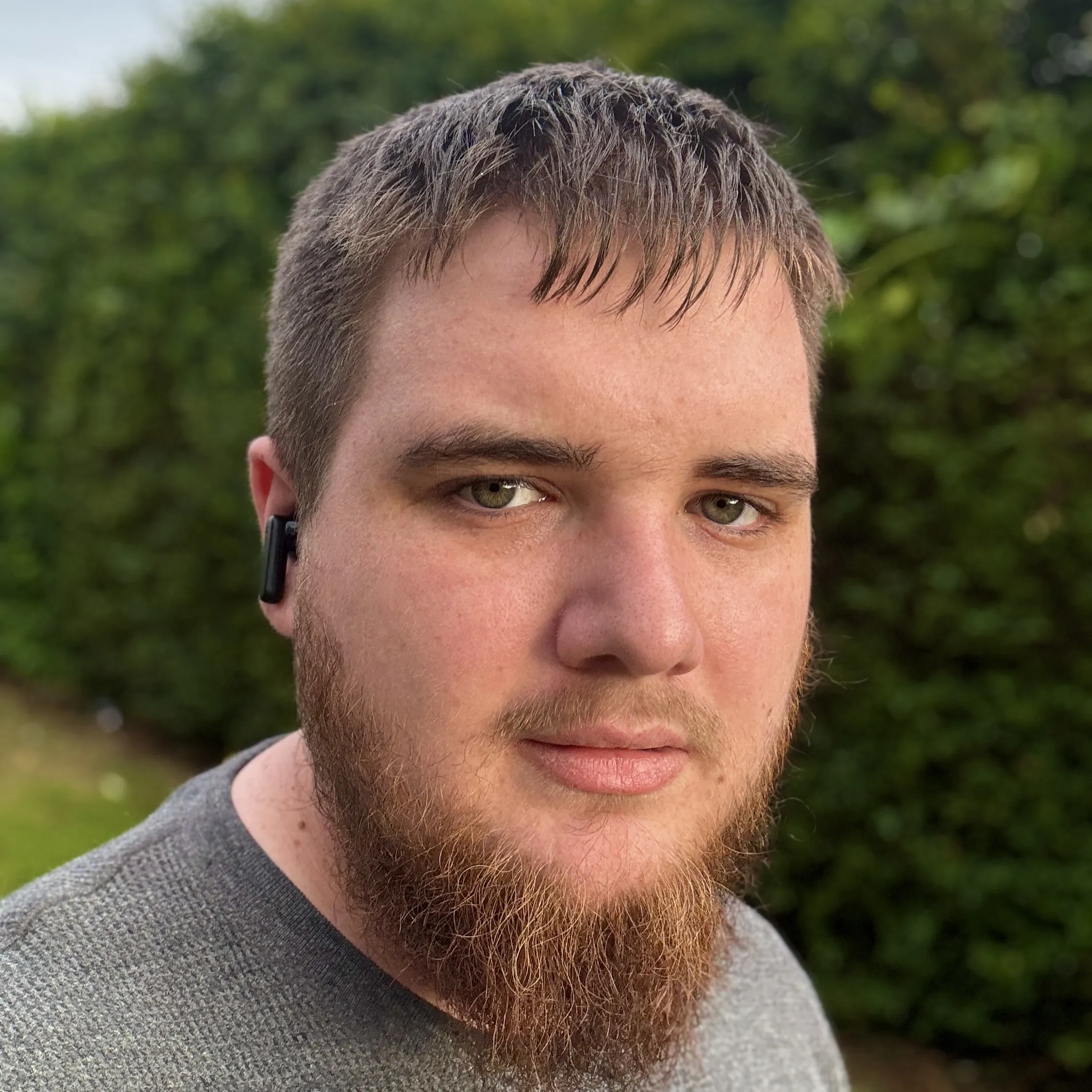 Callum, founder of Hello Tech, standing outdoors with a calm expression, short brown hair, a beard, and wearing a grey sweater and wireless earbuds. A green hedge is visible in the background.